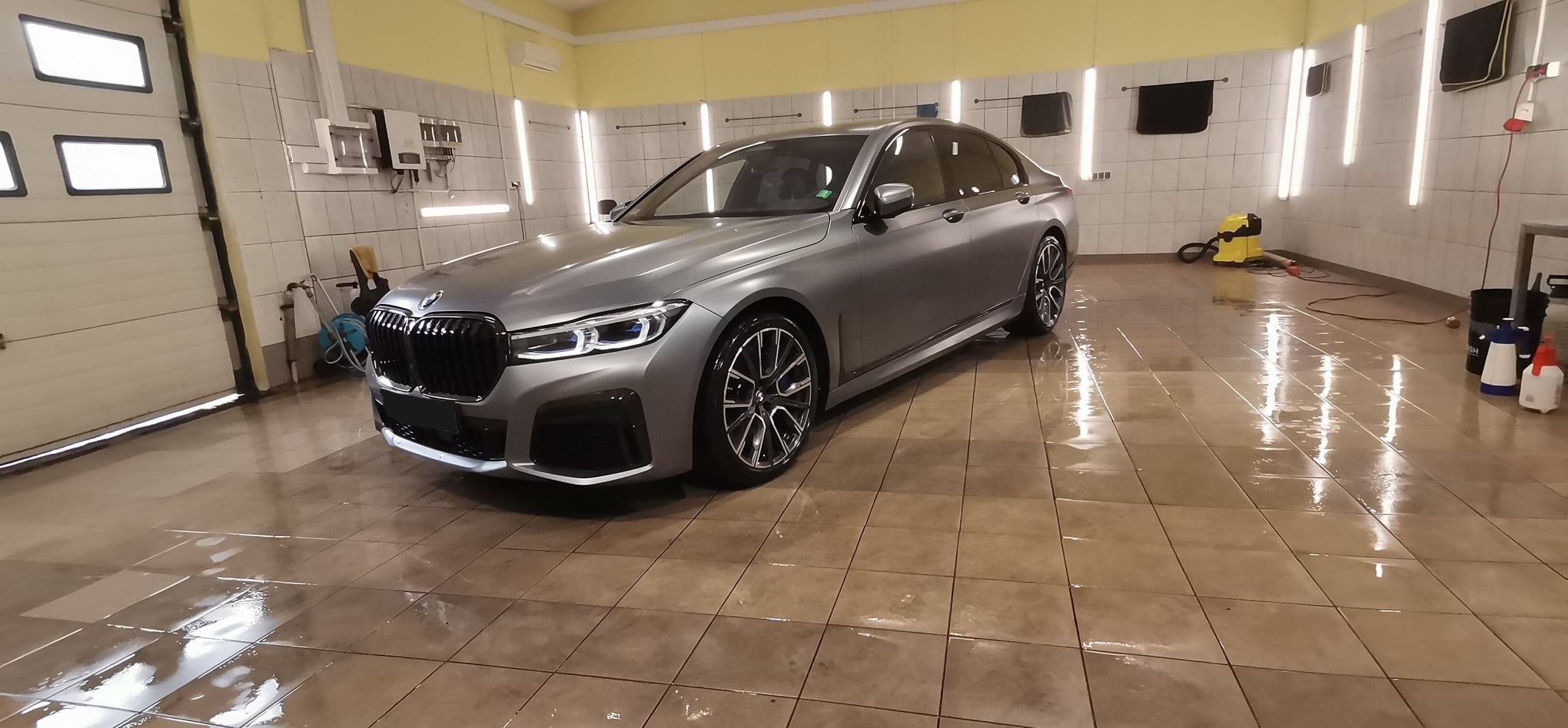 Szary sedan BMW serii 7 po myciu w profesjonalnej myjni, widoczne mokre płytki podłogowe i elementy wyposażenia myjni.