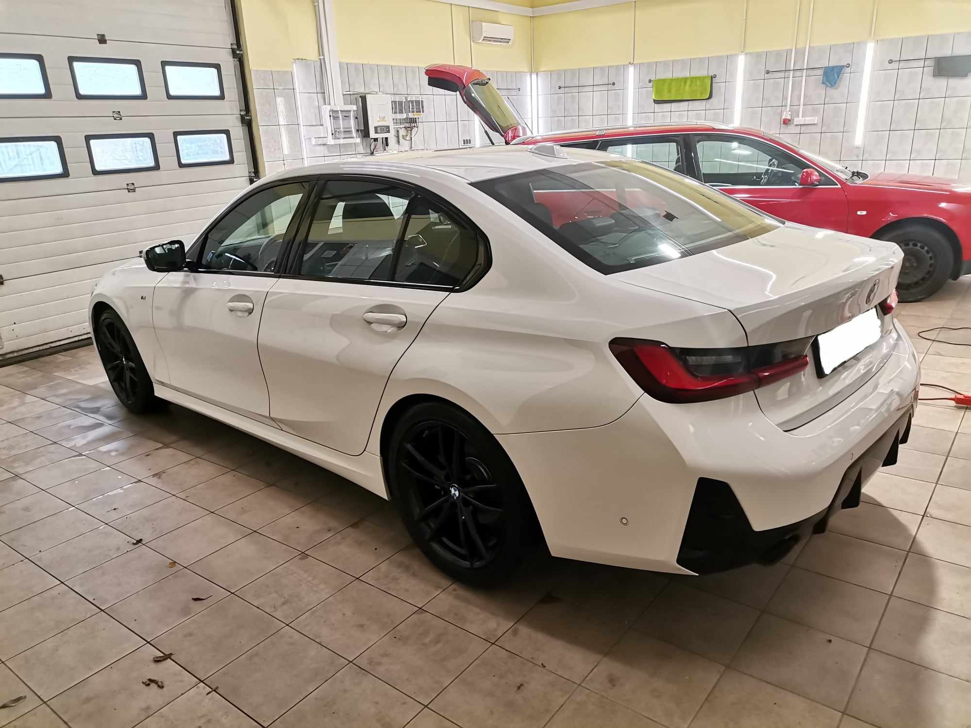Biały samochód BMW serii 3 na kafelkowanej podłodze w warsztacie, z widocznym czerwonym samochodem w tle i otwartą klapą bagażnika.