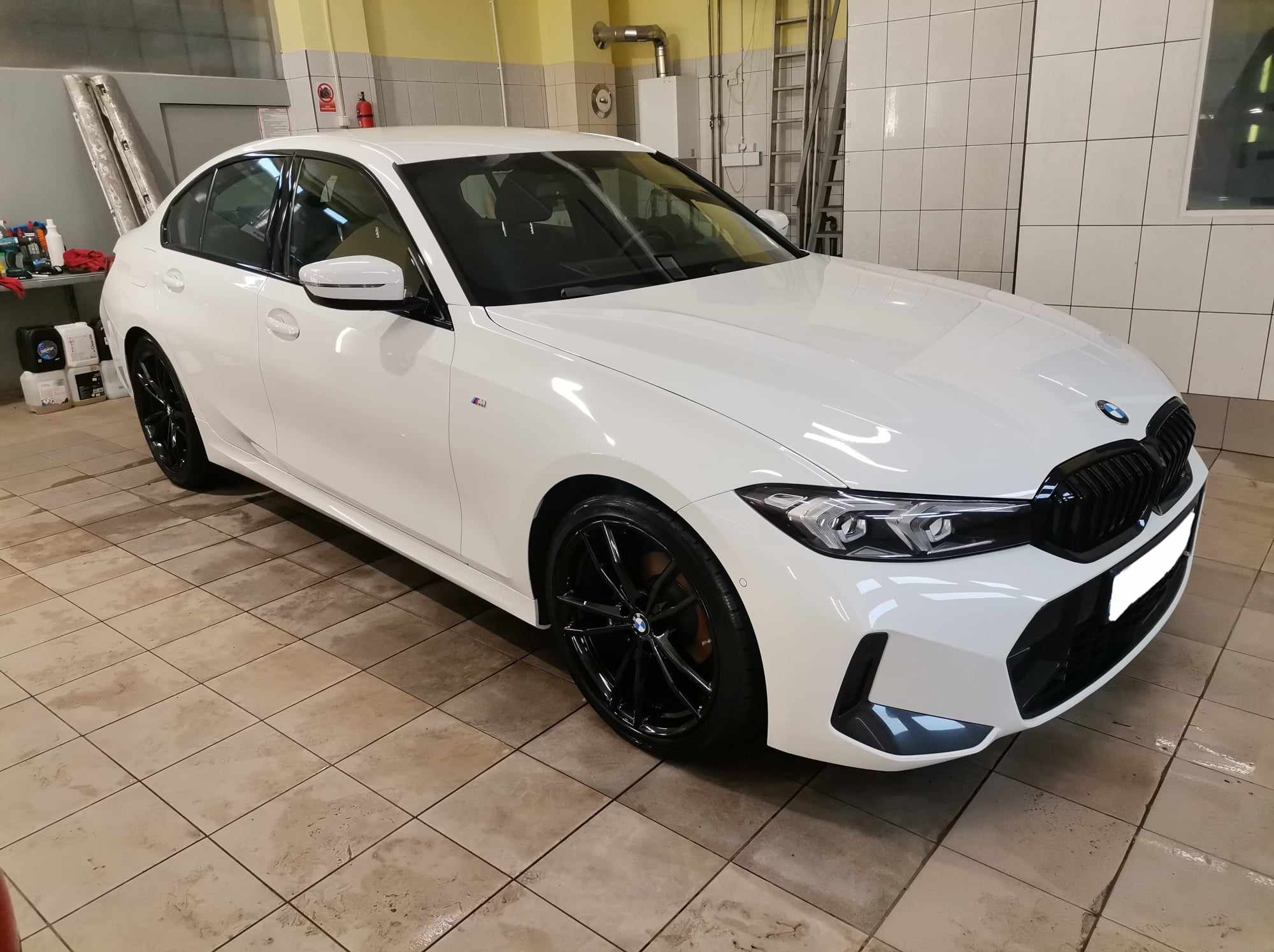 Białe BMW serii 3 po umyciu w myjni, z czarnymi felgami, widoczne na tle kafelkowanej ściany i podłogi myjni.