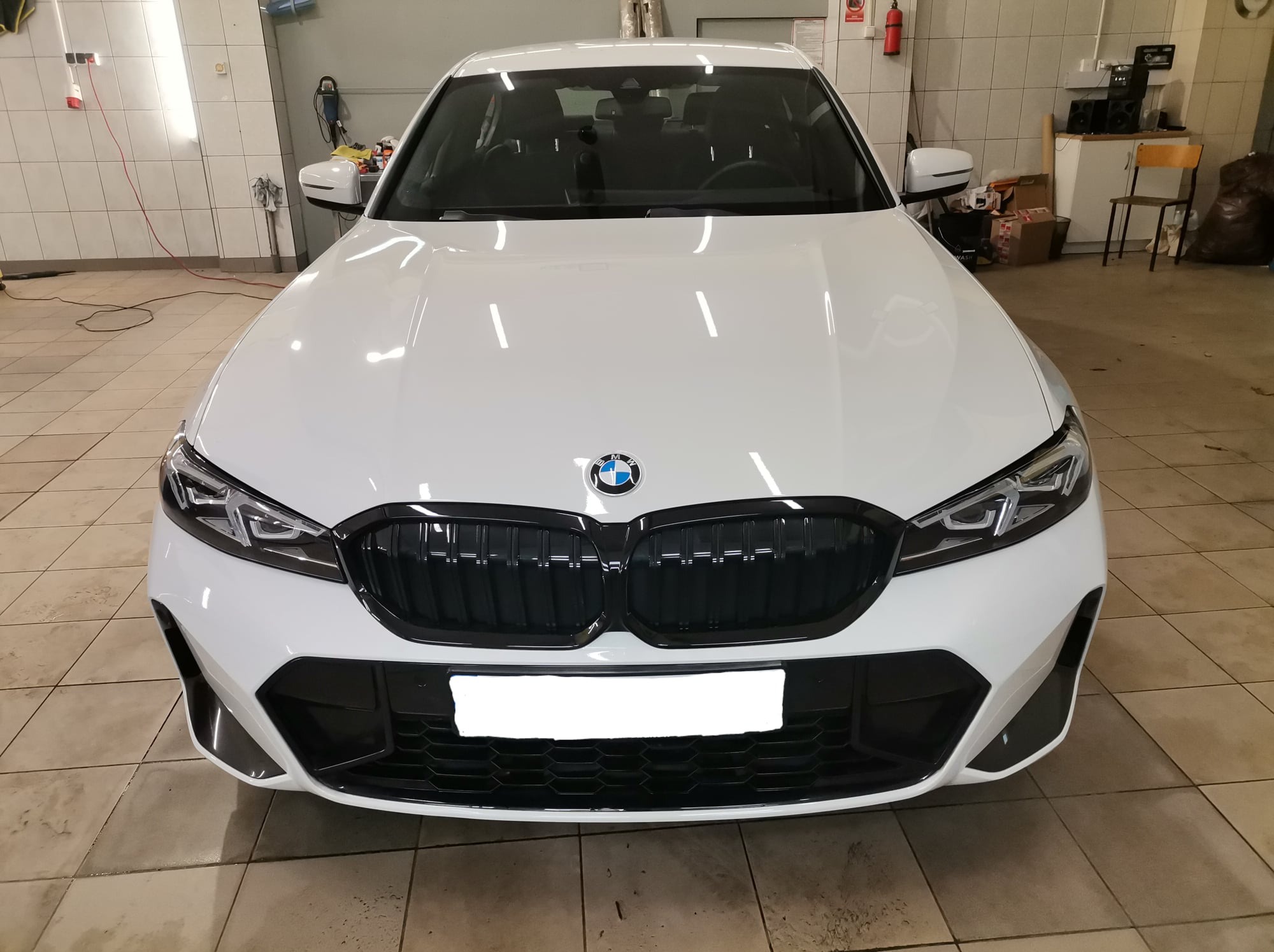 Biały samochód marki BMW, widok z przodu, po umyciu w myjni, z widocznymi kroplami wody na karoserii i czarnym grillem, na tle jasnych płytek podłogowych i ściany myjni.