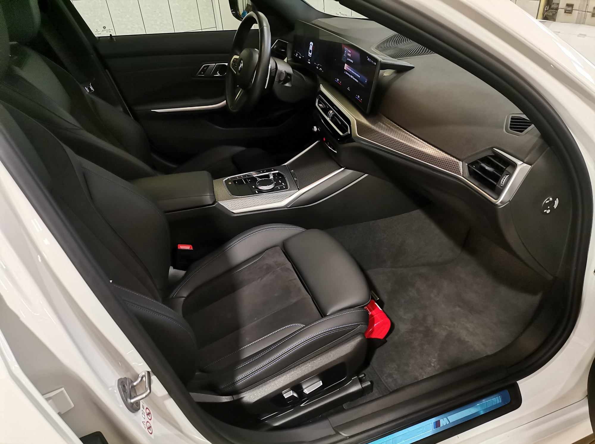 Wnętrze białego samochodu z czarną skórzaną tapicerką, widoczny fotel kierowcy, deska rozdzielcza z ekranem dotykowym i srebrnymi detalami, czerwony element pod siedzeniem.