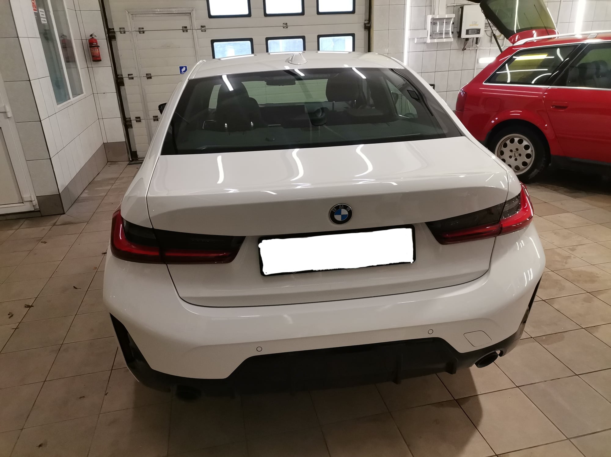 Tył białego samochodu BMW w hali z podnośnikiem i czerwonym Audi w tle, widoczne światła, tablica rejestracyjna zasłonięta, czujniki parkowania i pojedynczy wydech.