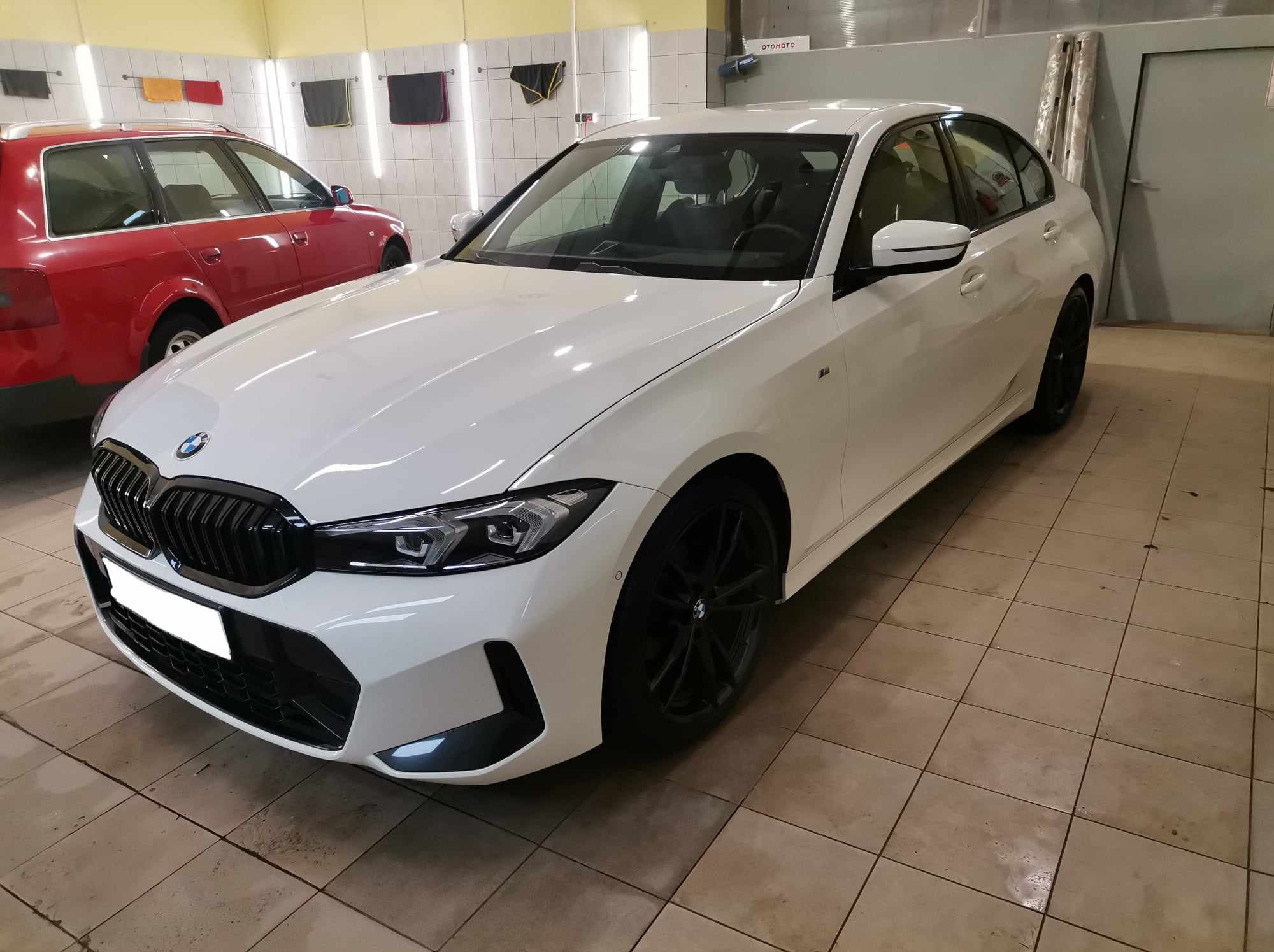 Białe BMW serii 3 po umyciu, widoczne na tle myjni z płytkami na podłodze i ręcznikami na ścianie, z czerwonym Audi w tle.