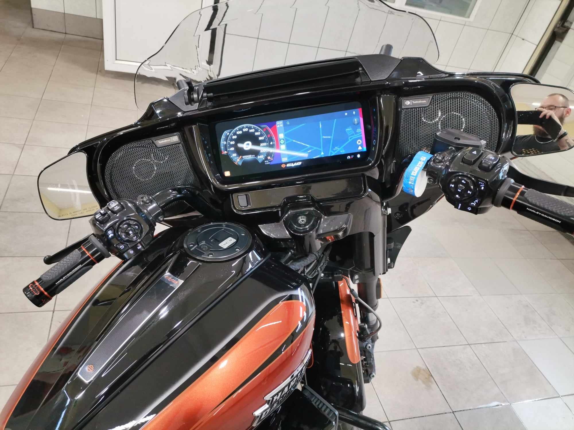Czarny motocykl Harley-Davidson z pomarańczowymi akcentami, widok z przodu na kierownicę z ekranem nawigacji i głośnikami, odblask osoby robiącej zdjęcie w lusterku.