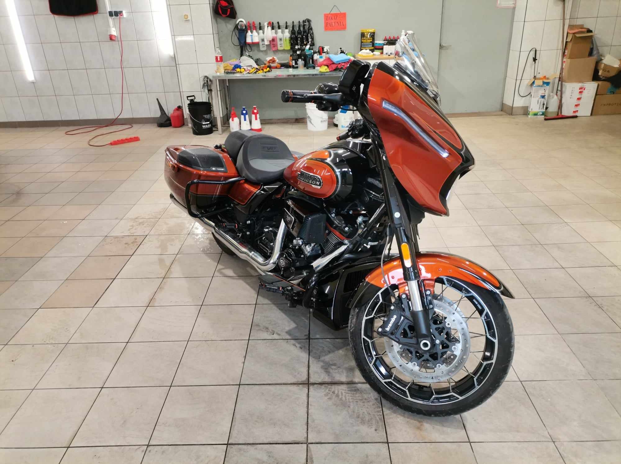 Lśniący, pomarańczowo-czarny motocykl Harley-Davidson po umyciu, zaparkowany na kafelkowanej podłodze myjni, z widocznymi butelkami środków czyszczących na stole w tle.
