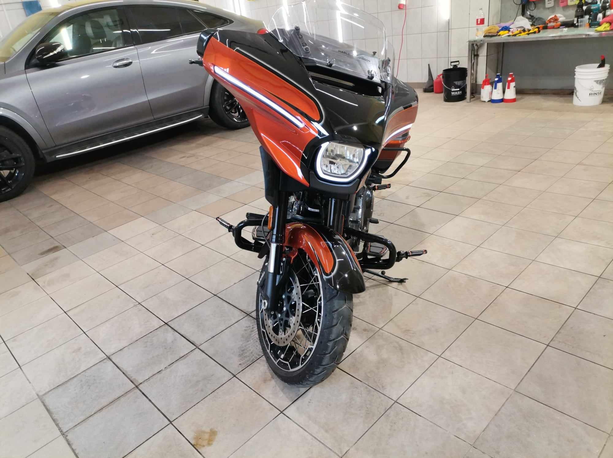 Pomarańczowo-czarny motocykl turystyczny Harley-Davidson w myjni, widok z przodu na tle szarego samochodu i półek z preparatami.