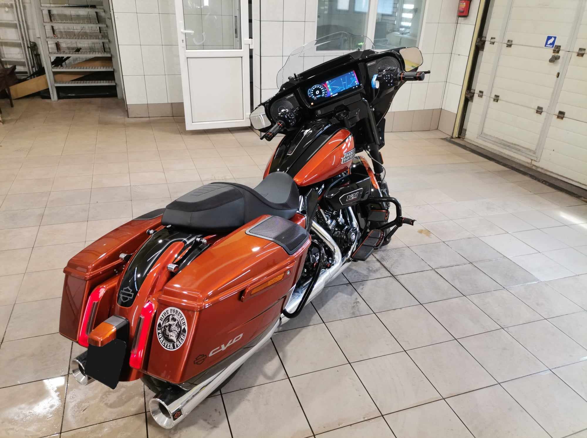 Pomarańczowo-czarny motocykl Harley-Davidson CVO w pomieszczeniu z białymi płytkami, widok z tyłu, z naklejką 'Ride Forever Free' na kufrze.