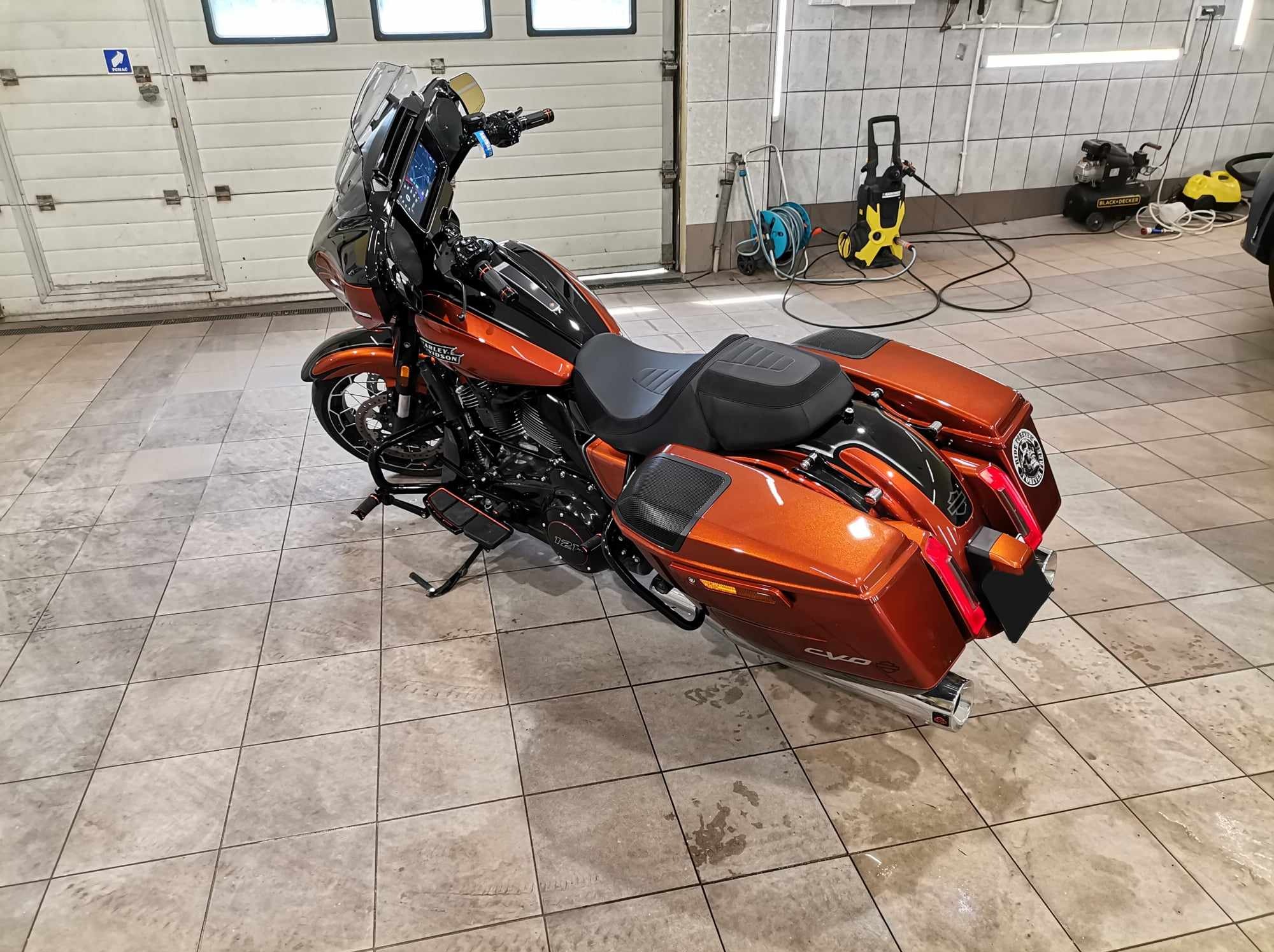 Pomarańczowo-czarny motocykl Harley-Davidson po umyciu, widoczny w garażu z myjką ciśnieniową w tle, na mokrej, kafelkowanej podłodze.