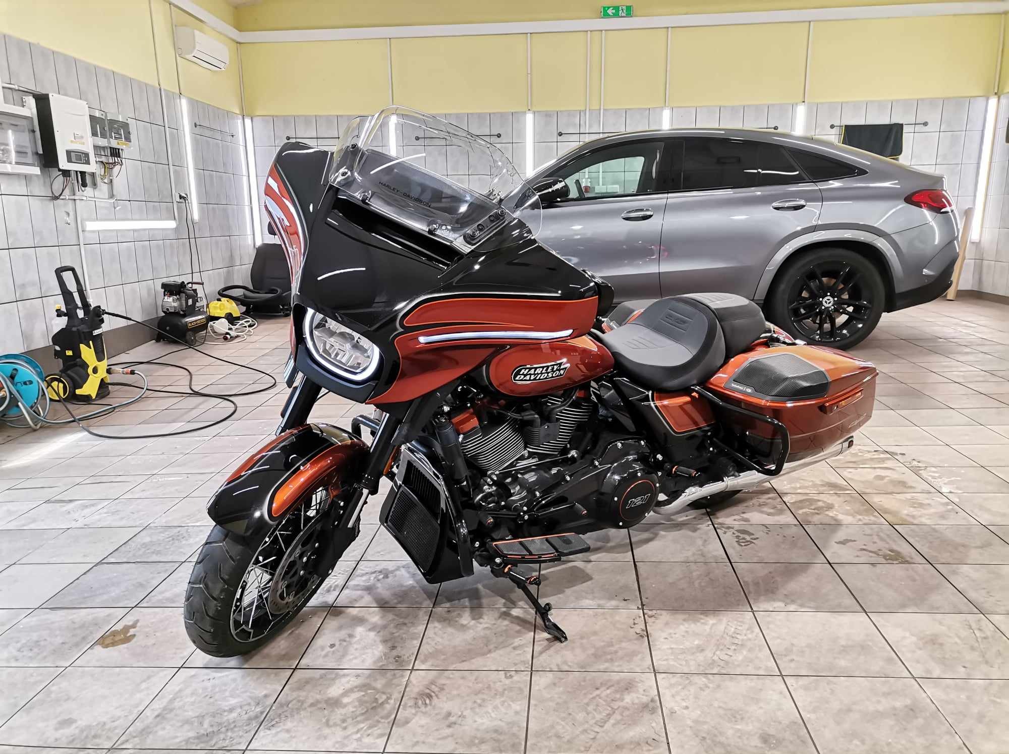 Pomarańczowo-czarny motocykl Harley-Davidson po umyciu w myjni, z widocznym samochodem w tle i sprzętem do mycia ciśnieniowego w rogu, na kafelkowanej podłodze.