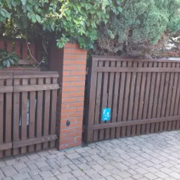 Brązowa drewniana brama z pionowych listew, osadzona na ceglanym słupku, z zielenią w tle i niebieską tabliczką informacyjną na skrzydle.