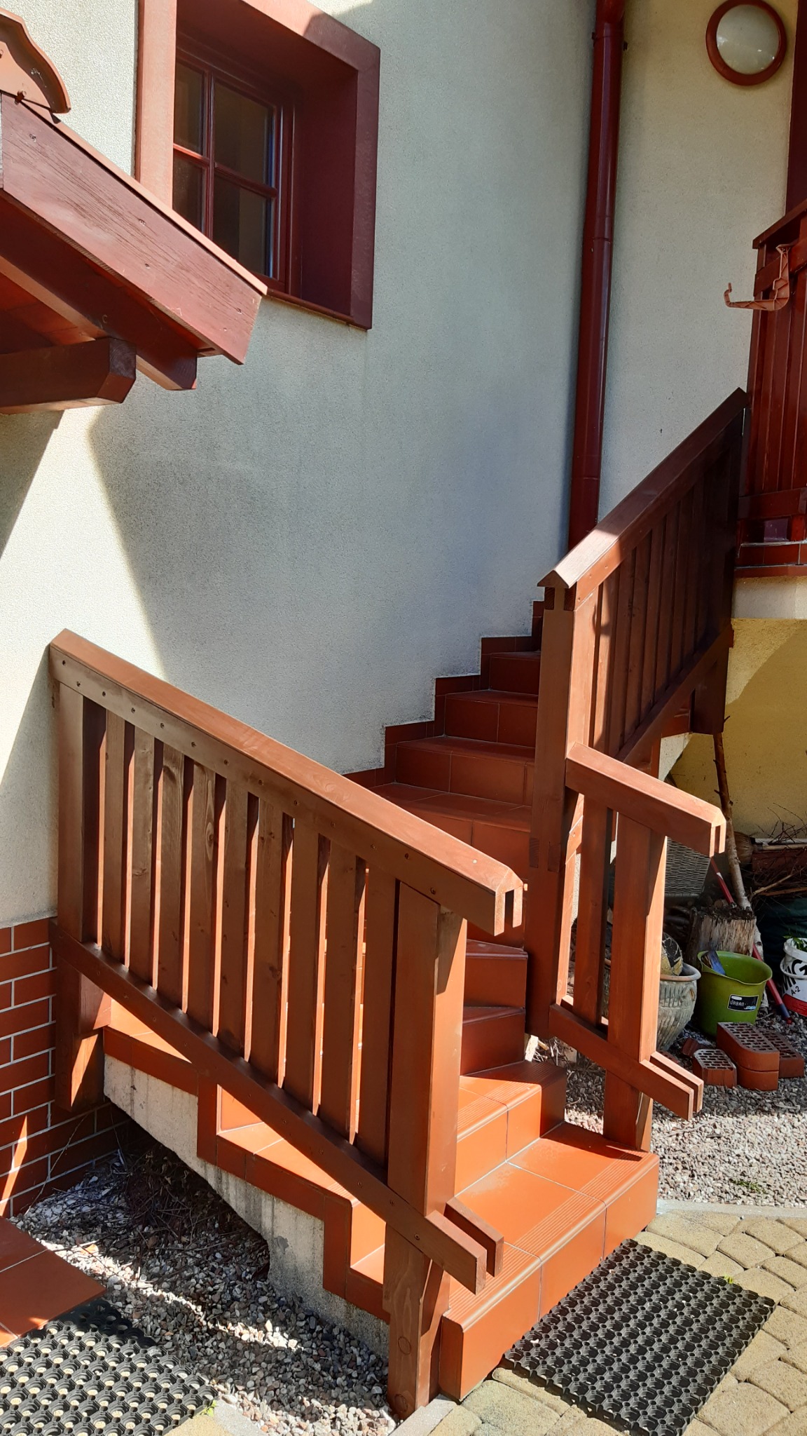 Zewnętrzne schody wyłożone terakotą z drewnianą balustradą, prowadzące do domu z oknem w czerwonej ramie i żółtą ścianą, widoczne doniczki i narzędzia ogrodnicze.