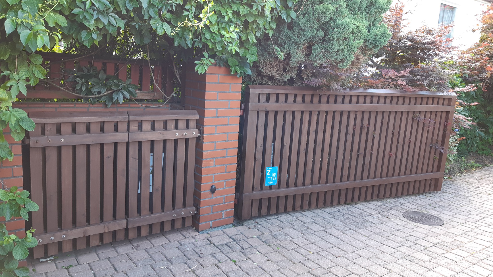 Brązowa drewniana brama z pionowych listew, osadzona na ceglanym słupku, z zielenią w tle i niebieską tabliczką informacyjną na skrzydle.