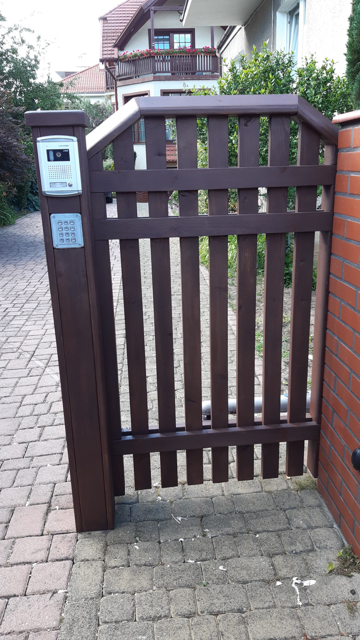 Ciemnobrązowa drewniana furtka z domofonem i klawiaturą numeryczną zamontowana na słupku, prowadząca na posesję wyłożoną kostką brukową, widoczny fragment ceglanego muru i budynek w tle.