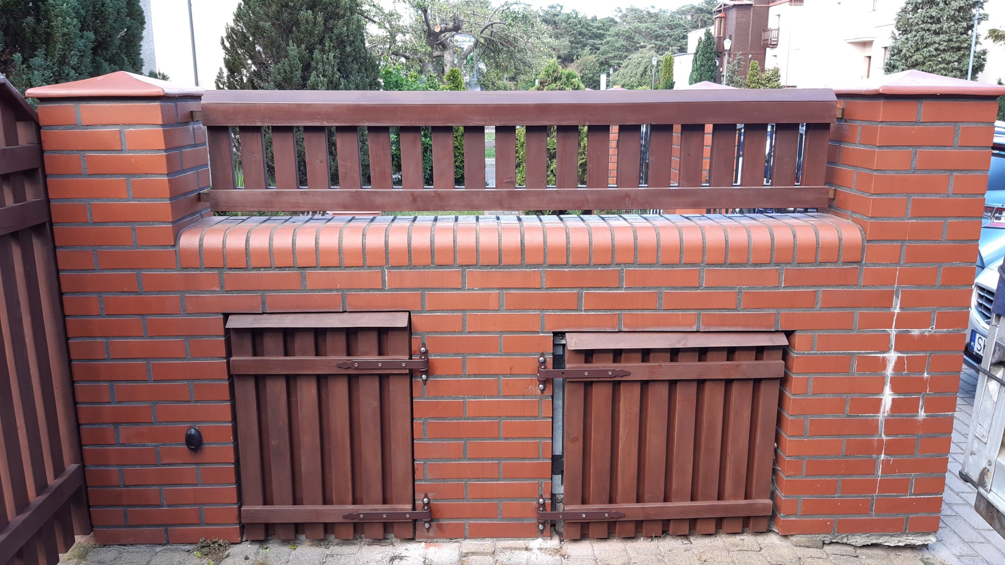 Mur z czerwonej cegły z drewnianym siedziskiem i dwoma małymi drewnianymi drzwiczkami na dole, tworzący element małej architektury ogrodowej.