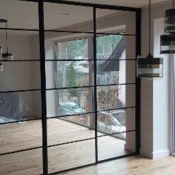 Nowoczesna, przeszklona ściana działowa z czarnymi szprosami, dzieląca przestrzeń salonu z widokiem na zewnątrz. W tle widoczny mebel zabezpieczony folią oraz minimalistyczne lampy wiszące...