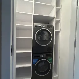 Biała szafa wnękowa z półkami, w której umieszczono pralkę i suszarkę marki Bosch w kolorze grafitowym.