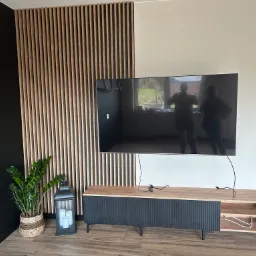 Salon z telewizorem wiszącym na ścianie z drewnianymi lamelami, szafka RTV z ryflowanymi frontami, roślina doniczkowa i lampion dekoracyjny.