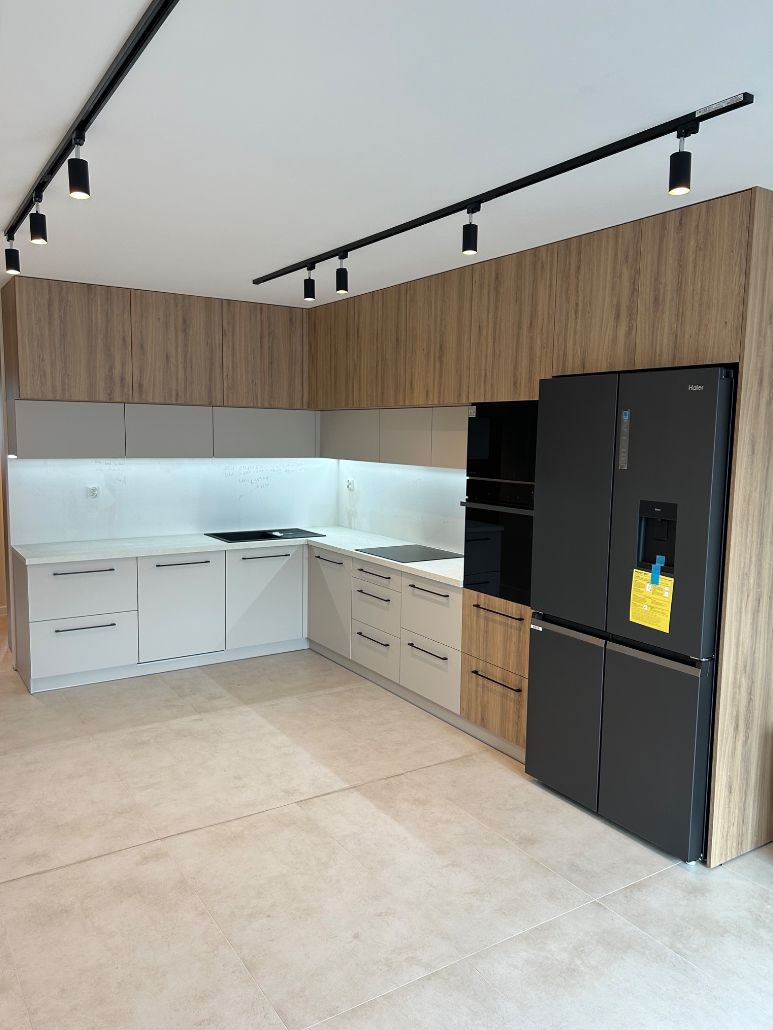 Nowoczesna kuchnia w odcieniach szarości i drewna z czarnym sprzętem AGD, oświetleniem szynowym i notatkami na ściance nad blatem.