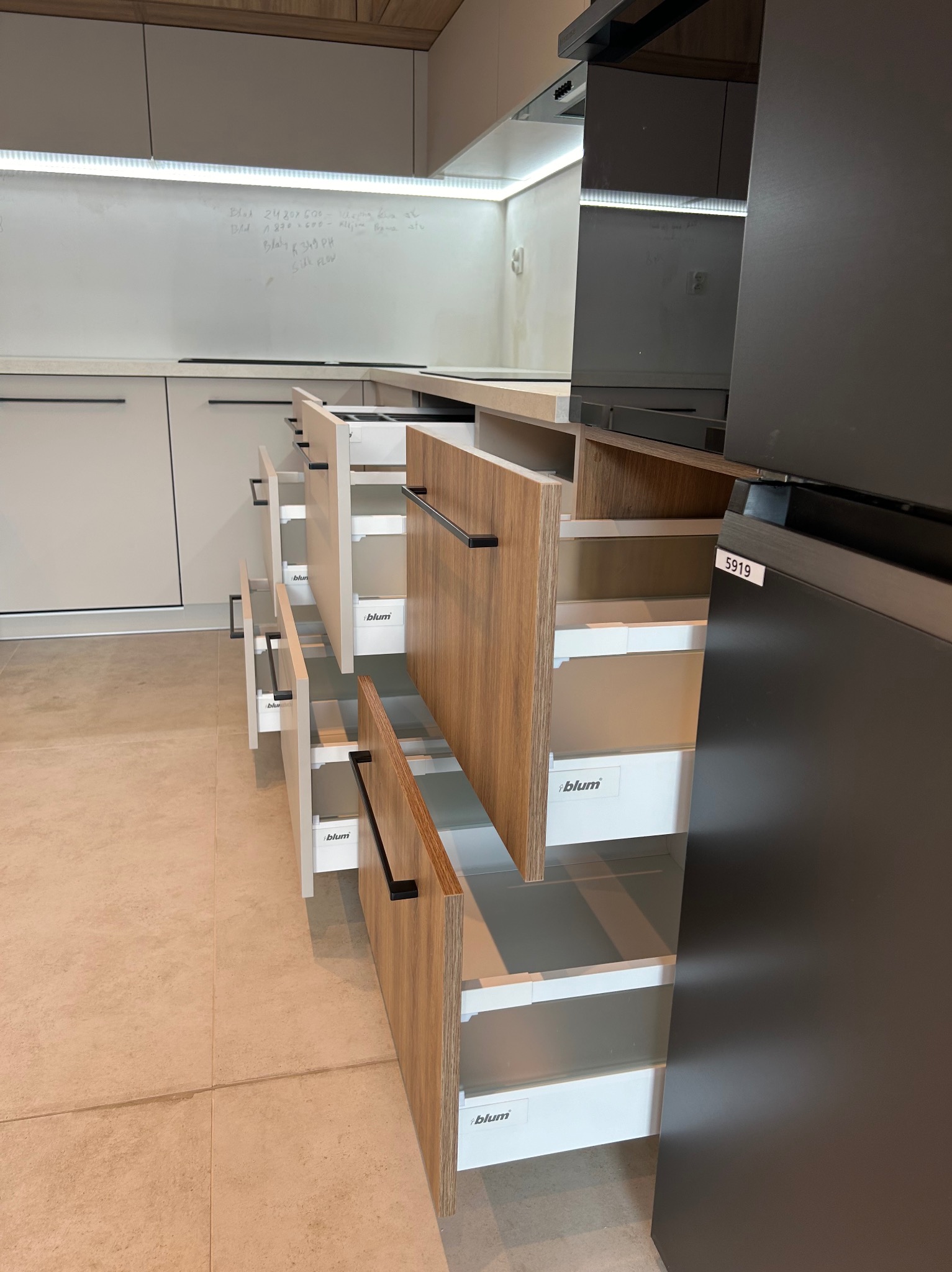 Otwarta szuflada kuchenna z drewnianym frontem i systemem Blum, widoczne białe, wewnętrzne przegródki, czarny uchwyt, w tle notatki na białej ścianie i fragment ciemnej zabudowy.