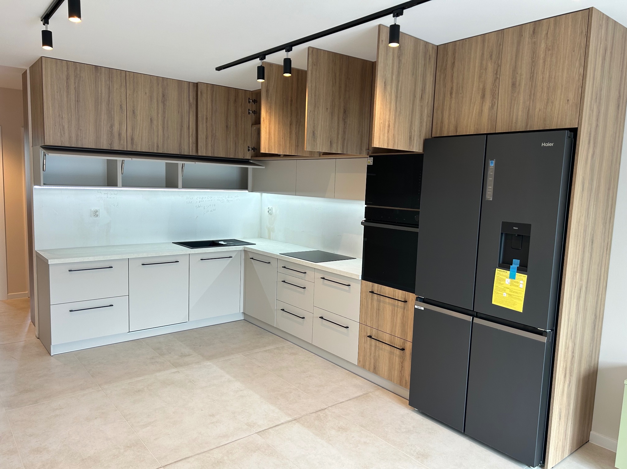 Nowoczesna kuchnia z frontami szafek w odcieniach bieli i drewna, czarnymi uchwytami i oświetleniem szynowym, uzupełniona o dużą, czarną lodówkę side-by-side.