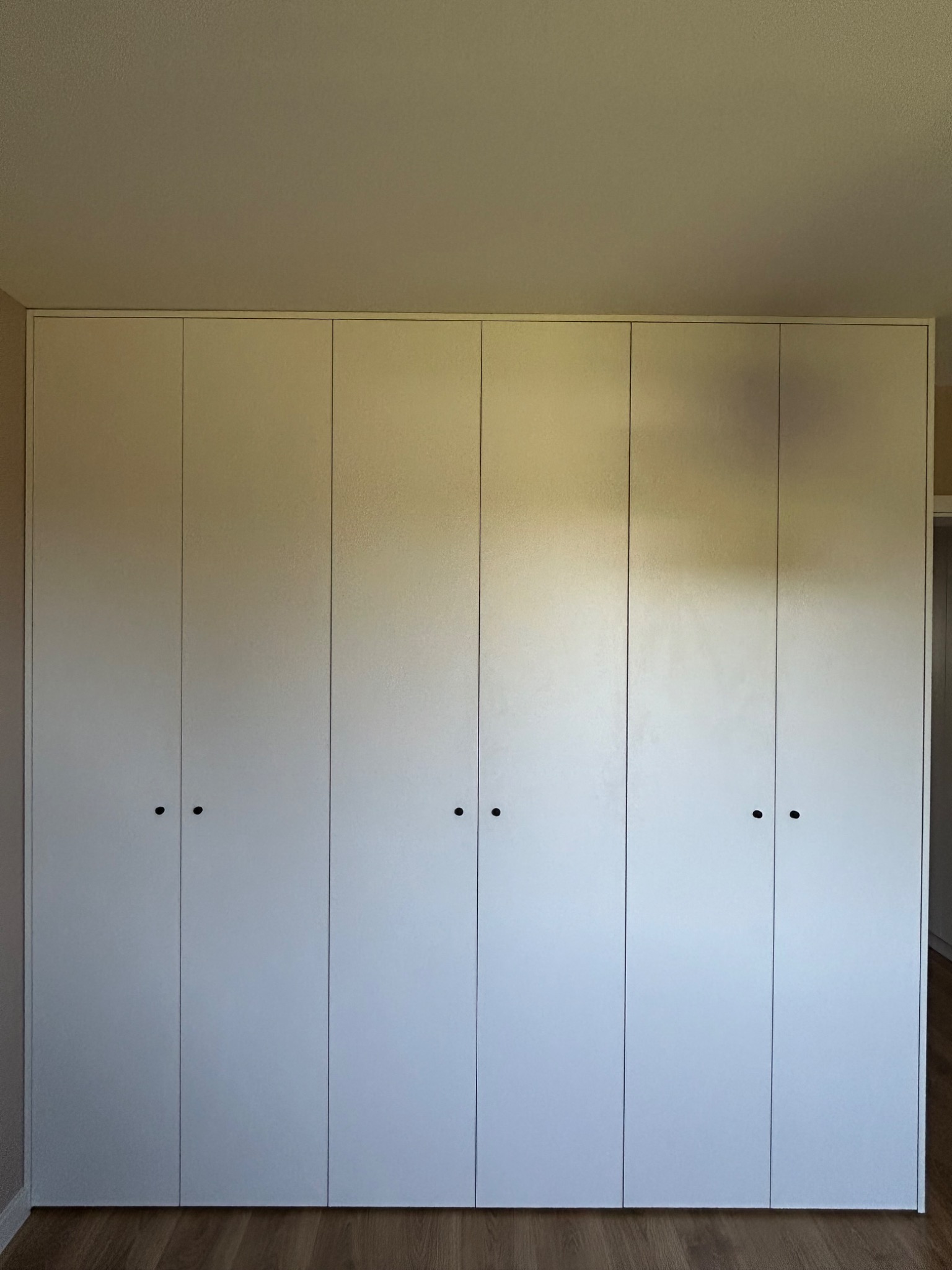 Biała szafa wnękowa z sześcioma drzwiami o minimalistycznym designie, z drobnymi, okrągłymi uchwytami na tle drewnianej podłogi i beżowej ściany.