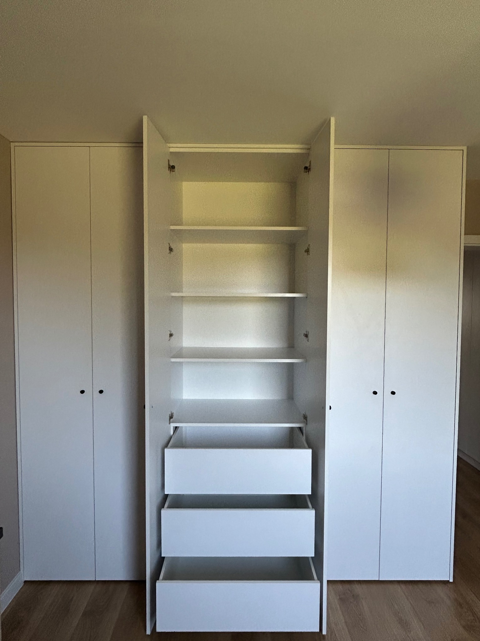 Biała szafa wnękowa z otwartymi drzwiami, ukazująca półki i trzy wysuwane szuflady, minimalistyczny design, jasne drewniane panele na podłodze.