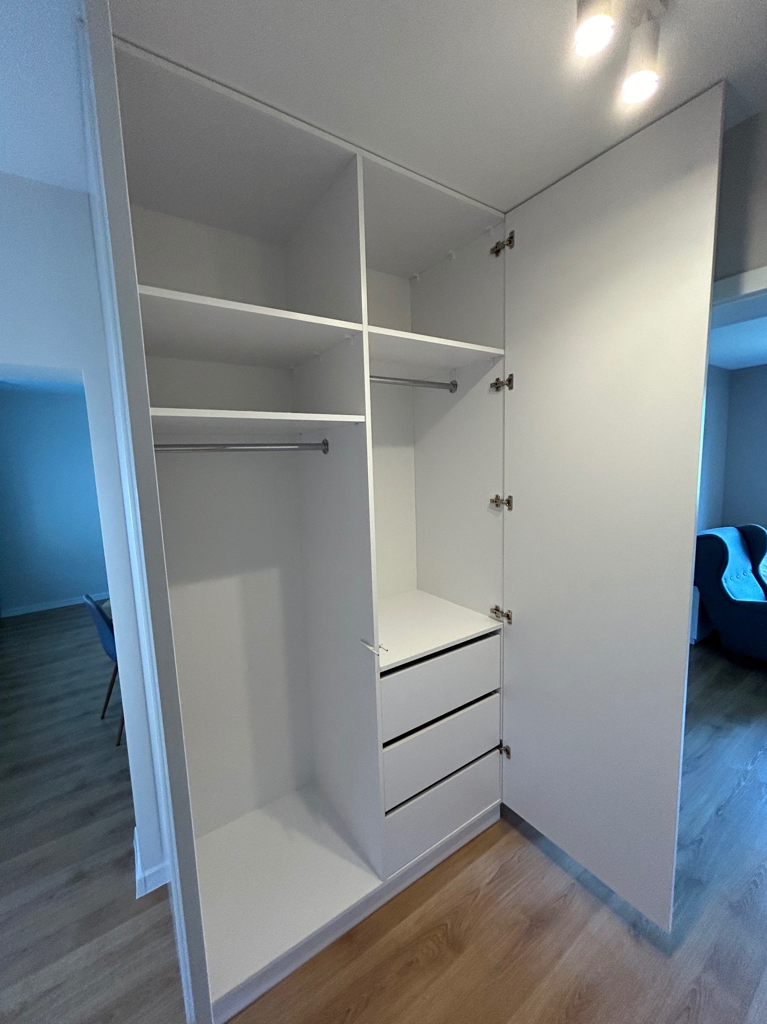 Wnętrze białej szafy wnękowej z półkami, drążkiem na ubrania i trzema szufladami, widok z otwartymi drzwiami, na podłodze panele imitujące drewno.