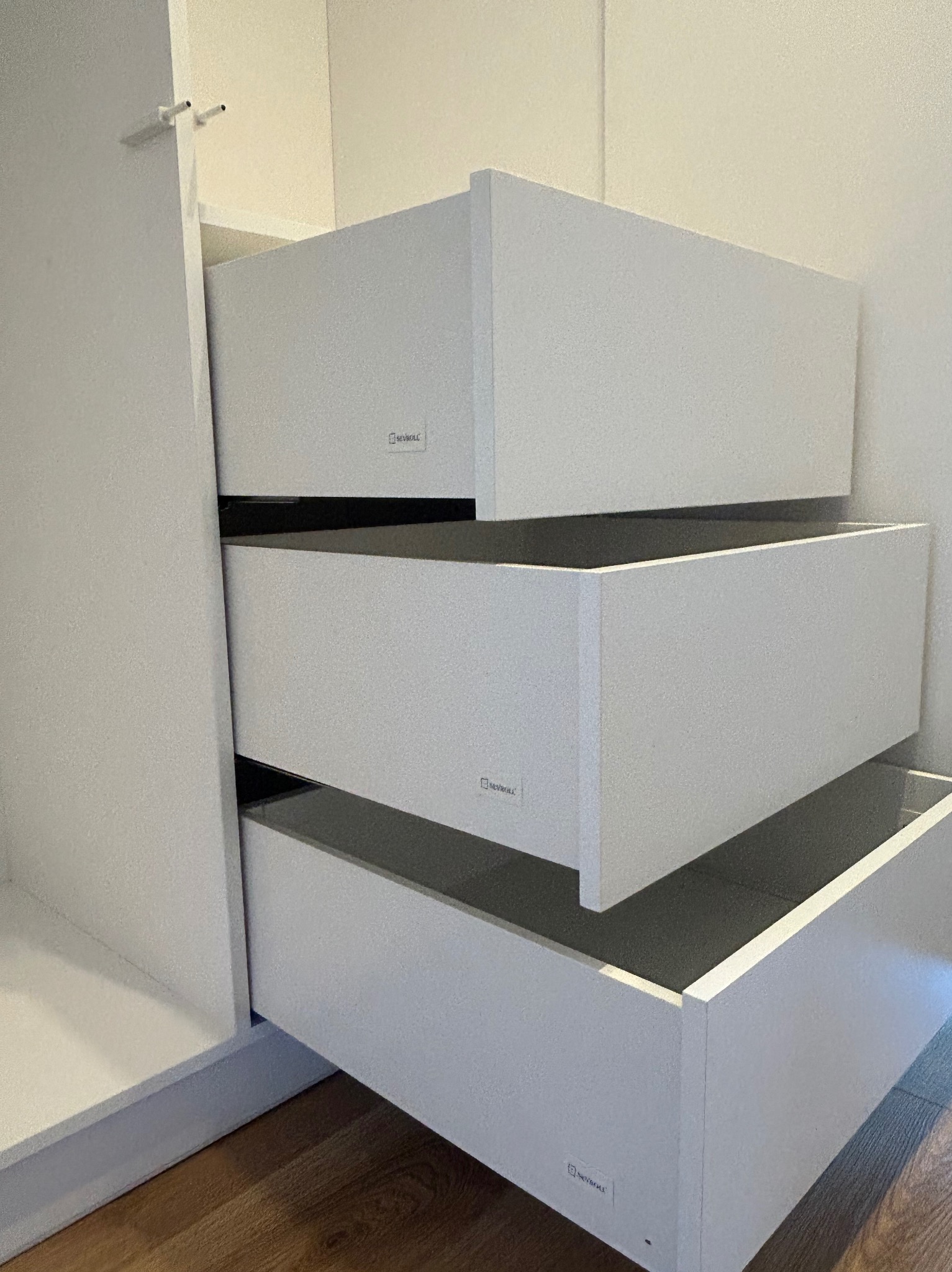 Wnętrze szafy wnękowej z trzema wysuwanymi, białymi szufladami z widocznym logo producenta na każdej z nich, na podłodze panele imitujące drewno.