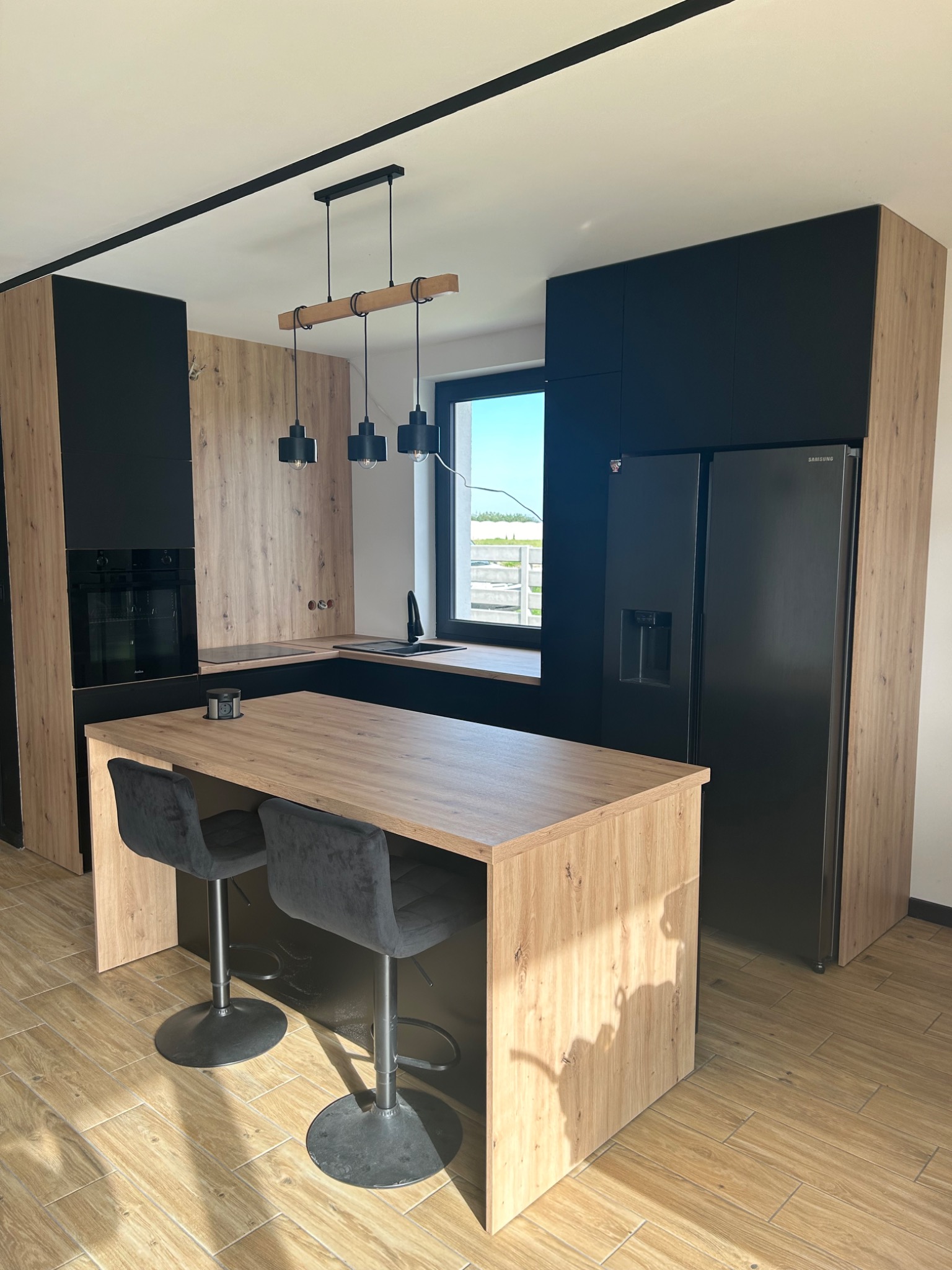 Nowoczesna kuchnia z wyspą i czarnymi akcentami, drewnopodobne fronty szafek, czarny sprzęt AGD, trzy czarne lampy wiszące nad wyspą, płytki podłogowe imitujące drewno, widok na zewnątrz przez okno.