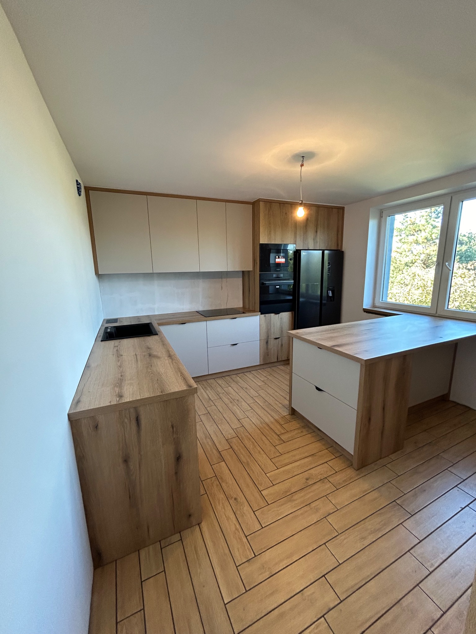 Jasna kuchnia z drewnianymi akcentami, widok na zabudowę kuchenną, wyspę z szufladami i czarny sprzęt AGD, podłoga wyłożona płytkami imitującymi drewno, oświetlenie punktowe.