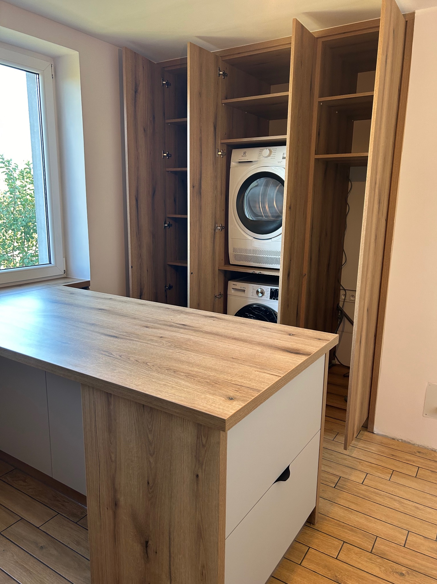 Szafa wnękowa z drewnianymi frontami, ukrywająca pralkę i suszarkę, z blatem roboczym i szufladami, widok na okno z zielenią za nim.