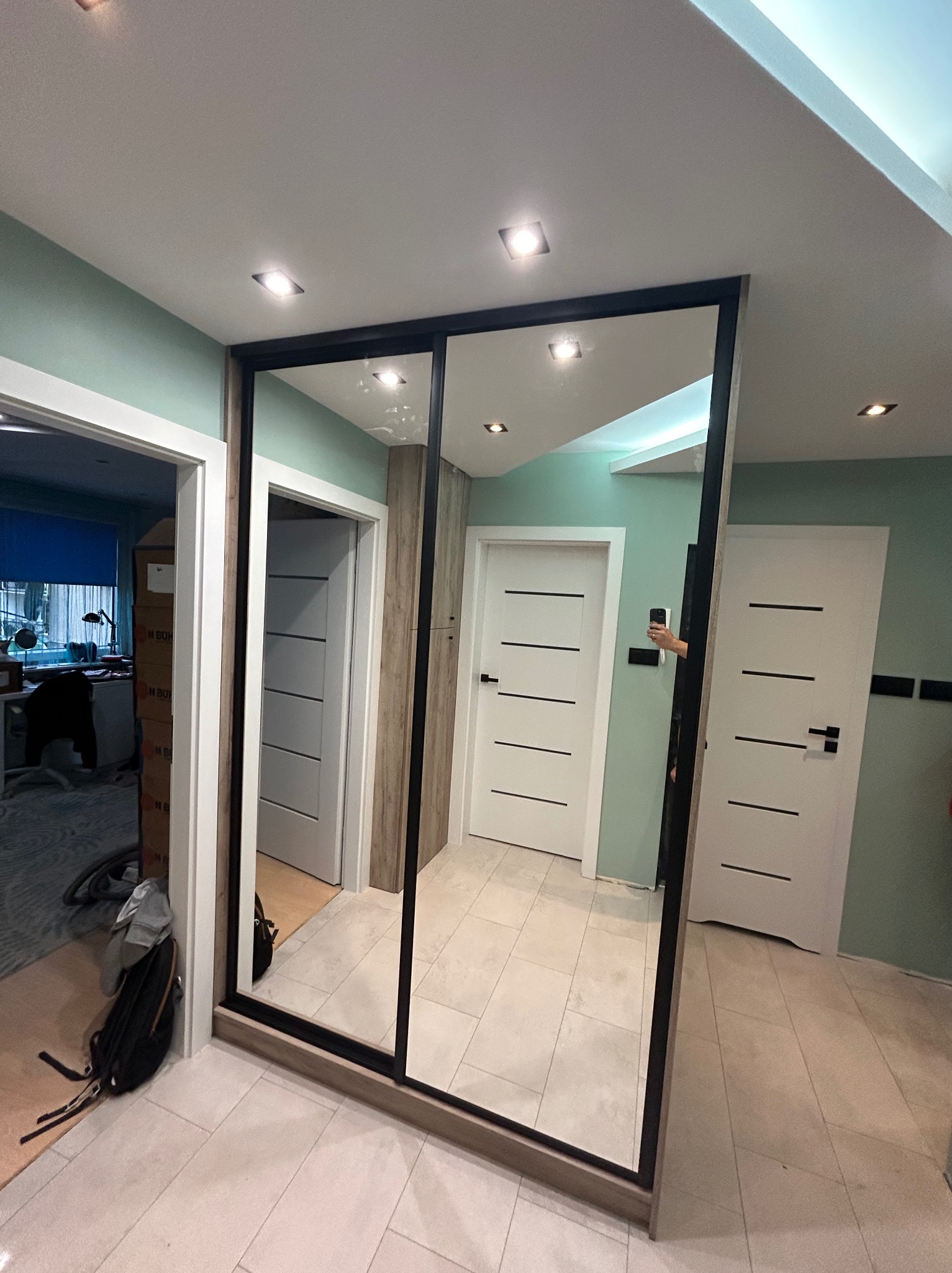 Dwu-drzwiowa szafa z lustrami w czarnej ramie, w tle widoczne drzwi wewnętrzne, jasna podłoga i ściany w odcieniach bieli i zieleni, w odbiciu widać rękę osoby robiącej zdjęcie.