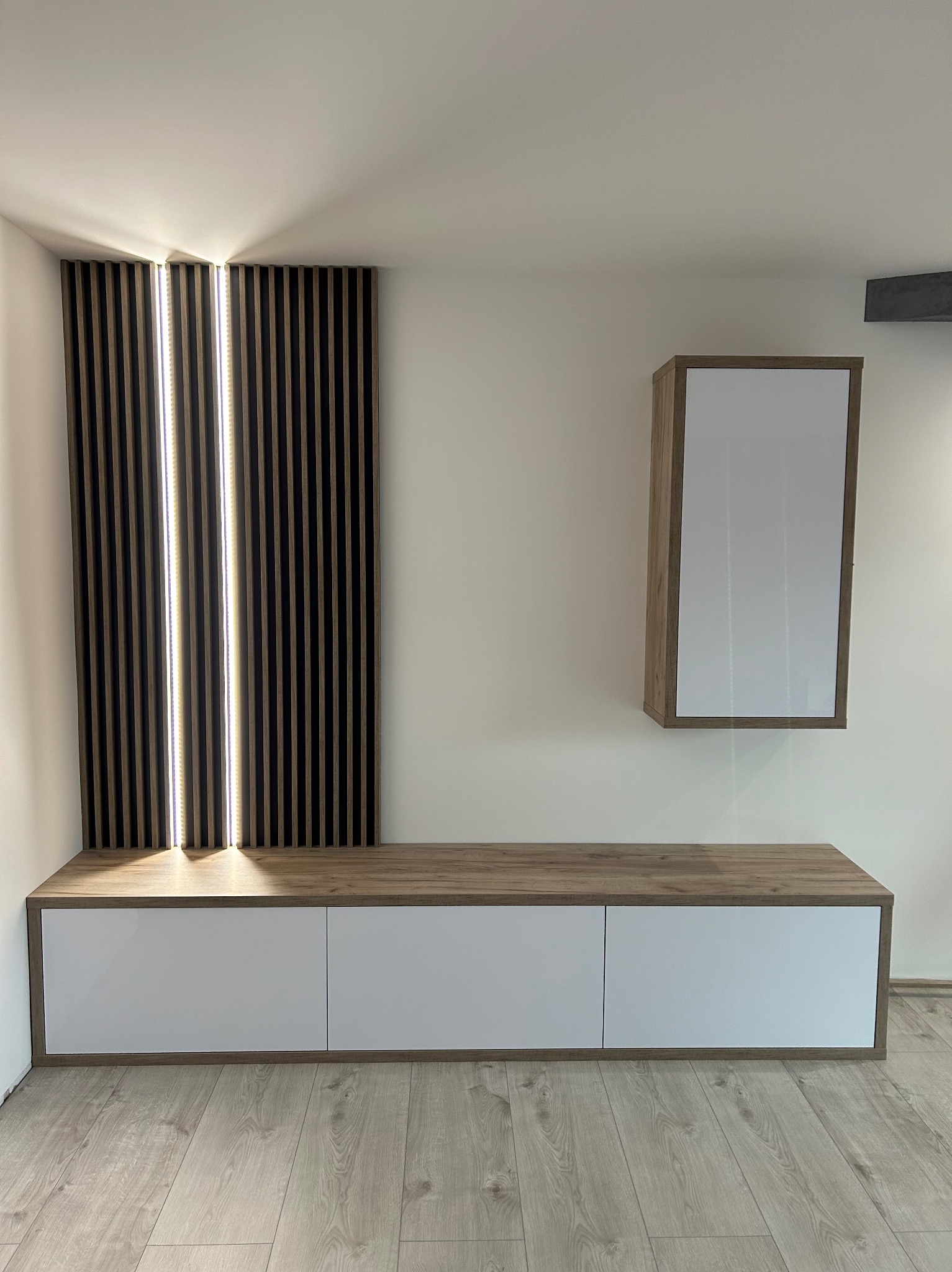 Nowoczesna aranżacja ściany z drewnianymi lamelami z podświetleniem LED, szafki RTV z białymi frontami i wiszącej szafki w kolorze drewna.