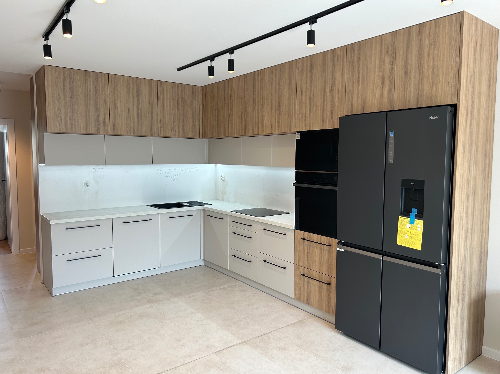 Nowoczesna kuchnia z jasnymi szafkami dolnymi i górnymi w odcieniach drewna, czarnymi uchwytami, oświetleniem szynowym oraz dużą, czarną lodówką typu side-by-side z żółtą kartką informacyjną.