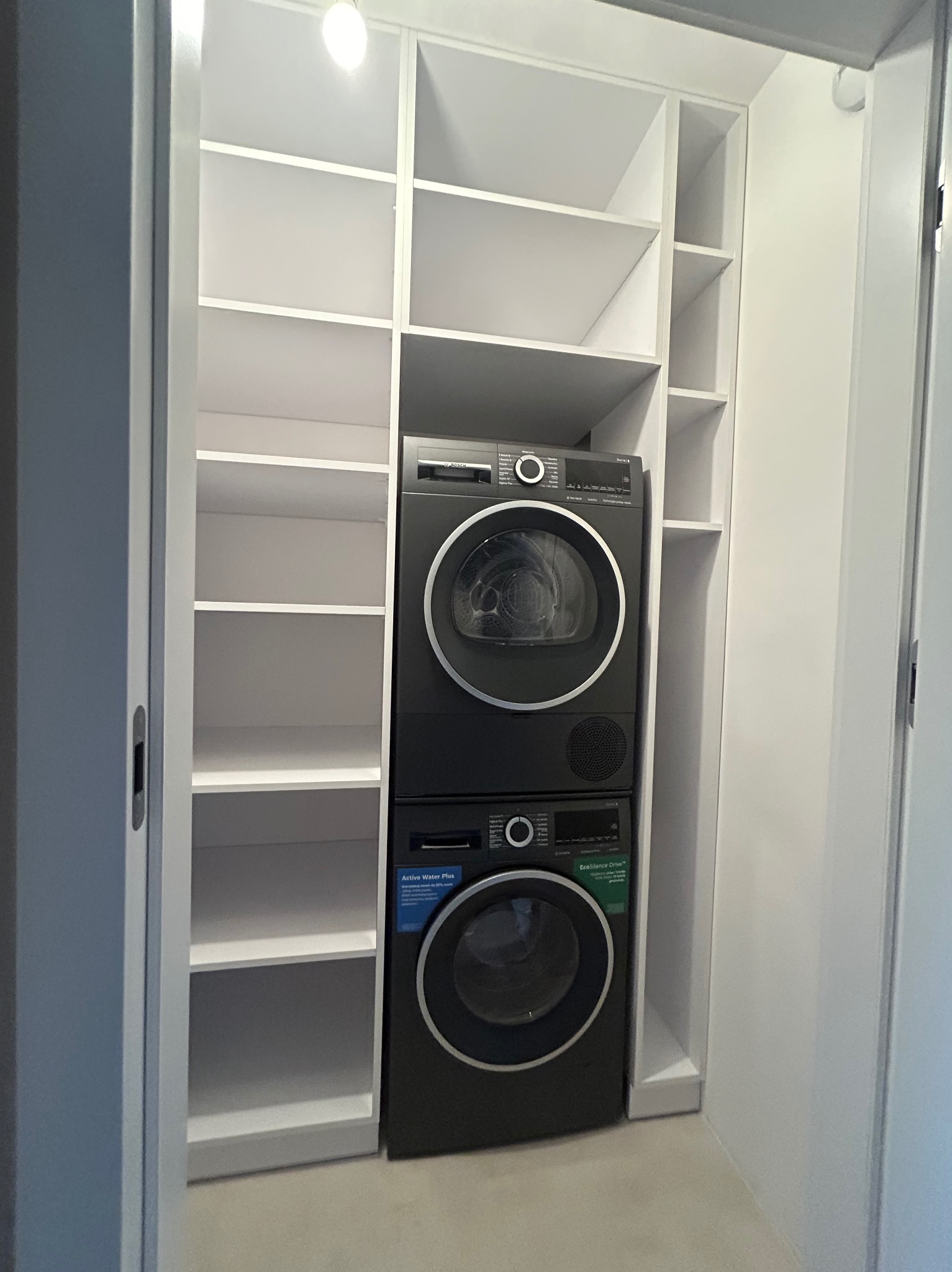Biała szafa wnękowa z półkami, w której umieszczono pralkę i suszarkę marki Bosch w kolorze grafitowym.