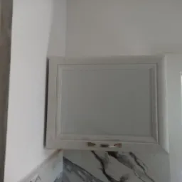 Górna szafka kuchenna w kolorze złamanej bieli z marmurowym blatem, widoczna częściowo przez futrynę drzwi, w tle salon z osobą siedzącą na kanapie.