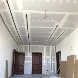 Montaż płyt karton gips na suficie z widocznymi przewodami elektrycznymi i trzema parami ciemnych drzwi w tle.