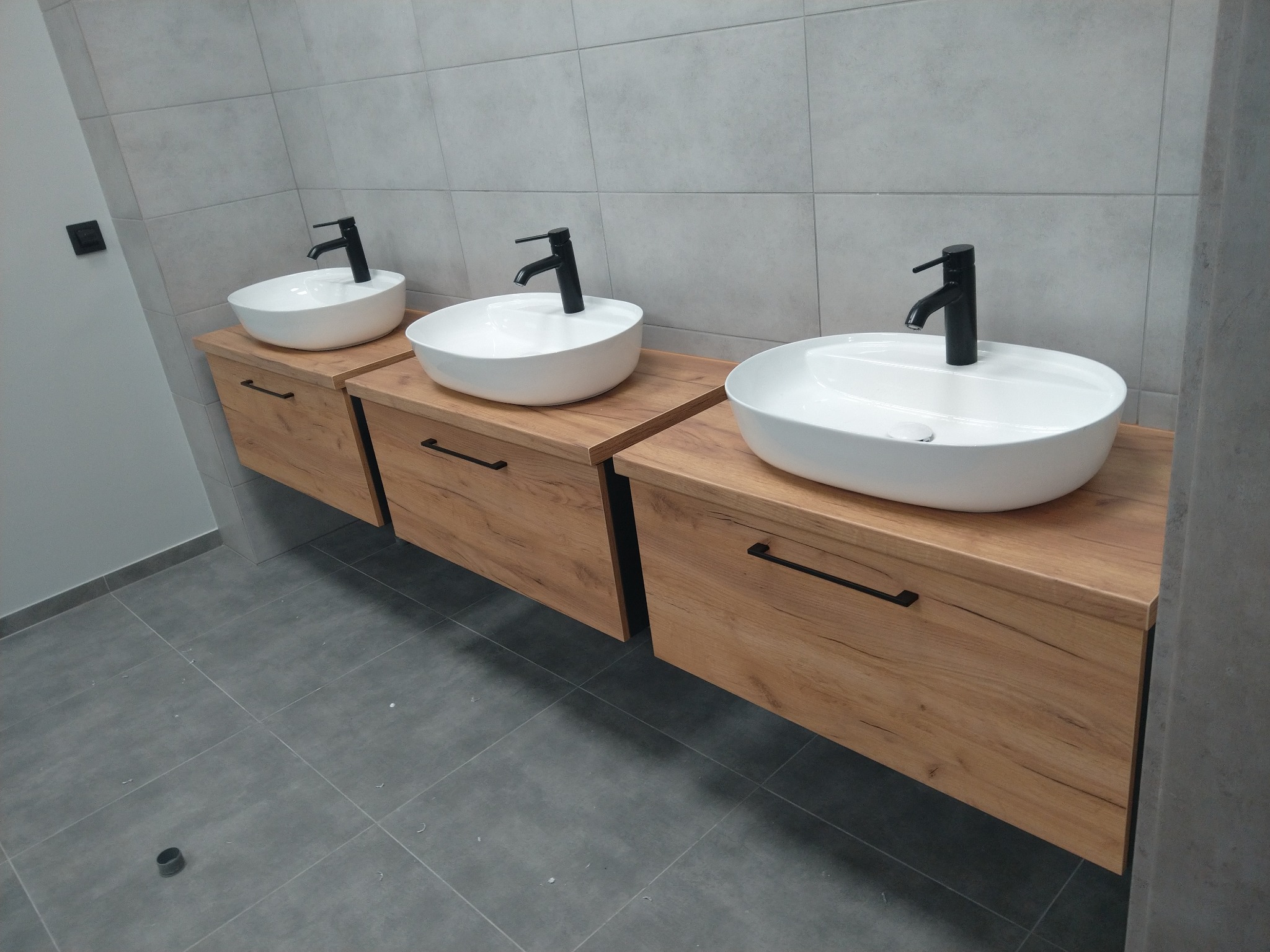 Trzy białe umywalki nablatowe z czarnymi bateriami zamontowane na drewnianych szafkach z poziomymi uchwytami w łazience z szarą glazurą i podłogą.