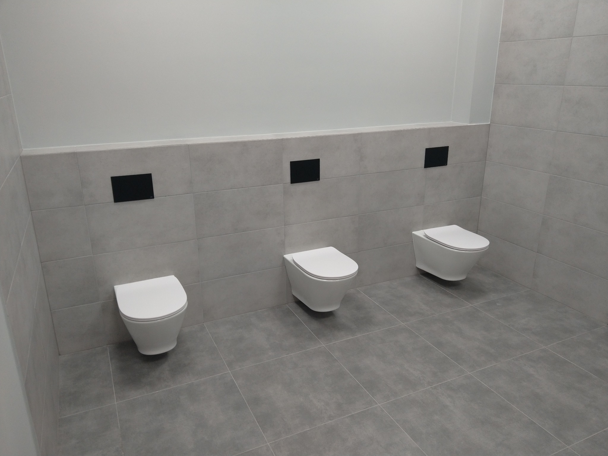 Trzy podwieszane toalety z czarnymi przyciskami spłukującymi zamontowane na szarej, kafelkowanej ścianie i podłodze w nowoczesnej łazience, widok z przodu.