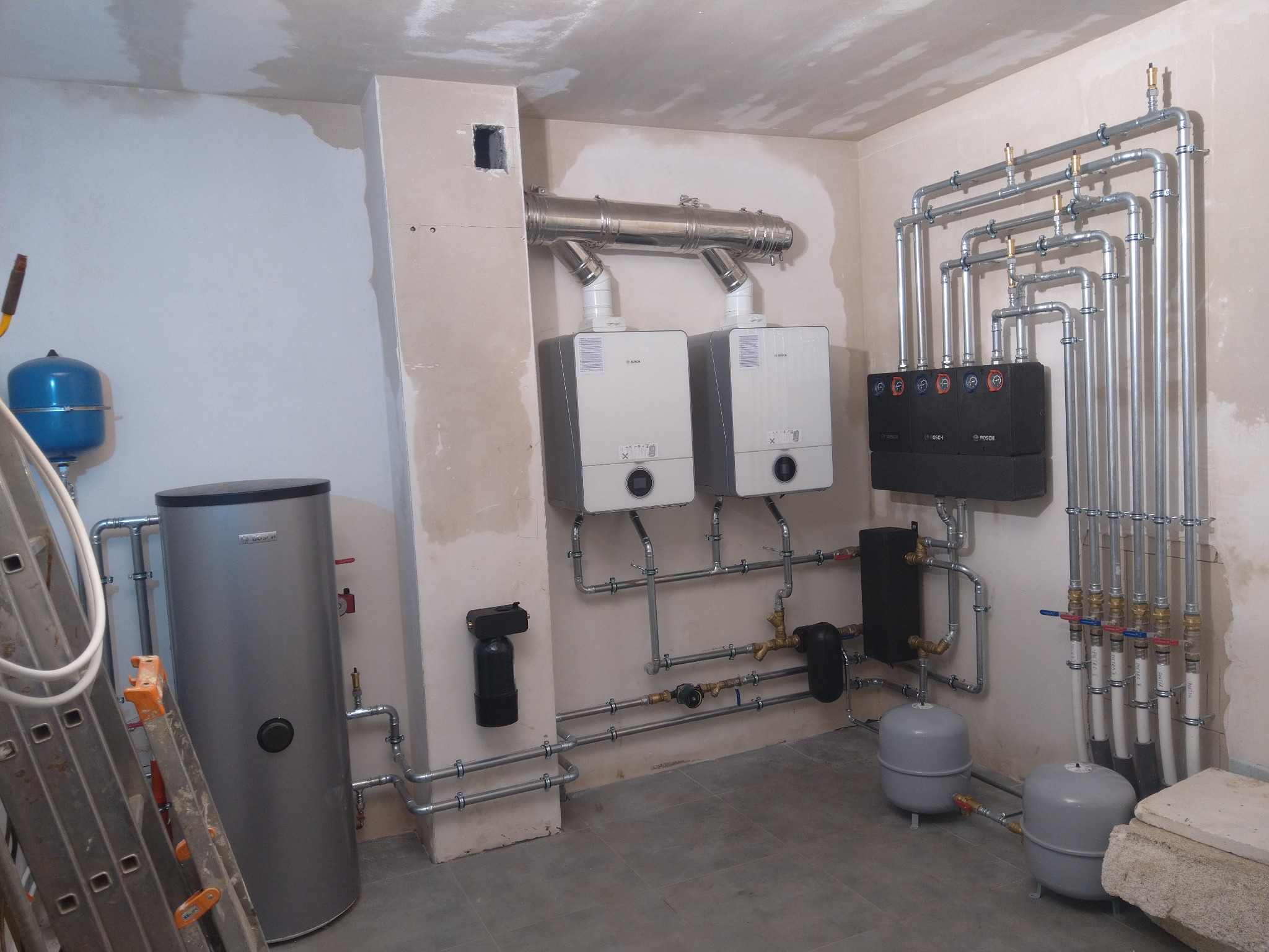 Dwa wiszące kotły gazowe z podłączonymi rurami spalinowymi i instalacją hydrauliczną, widoczny zasobnik ciepłej wody, rozdzielacz oraz zbiorniki wyrównawcze w surowym pomieszczeniu.