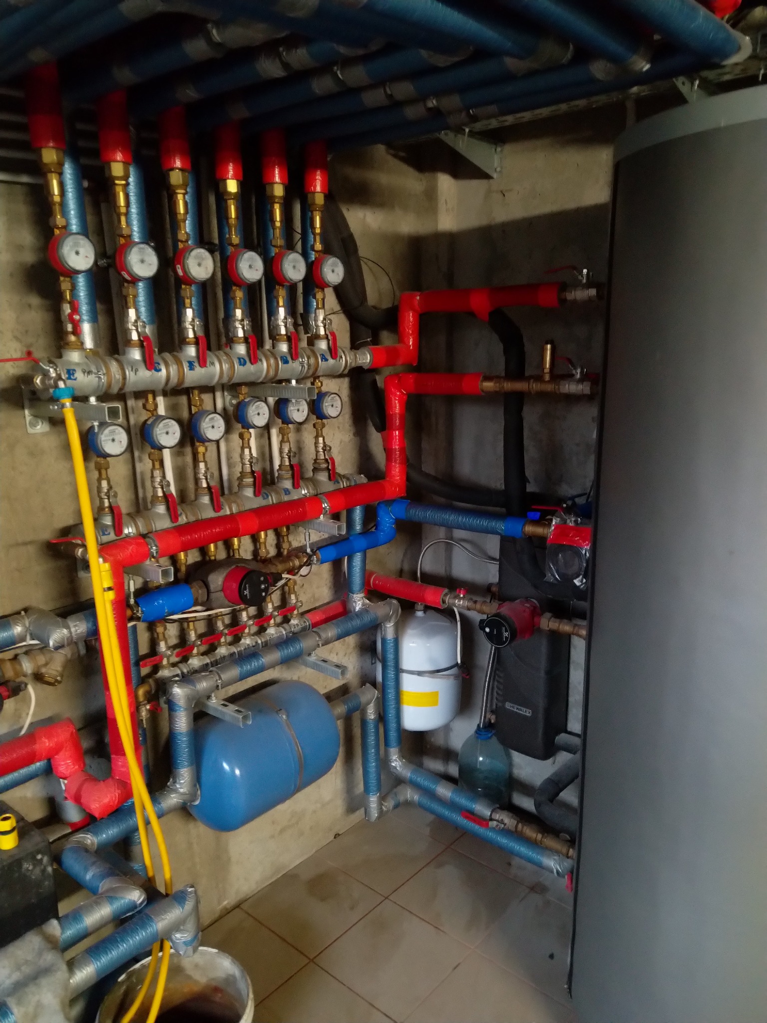 Rozbudowana instalacja hydrauliczna i grzewcza z licznymi rurami w kolorach czerwonym i niebieskim, manometrami i zbiornikami wyrównawczymi w kotłowni, widoczne elementy z mosiądzu i zawory.