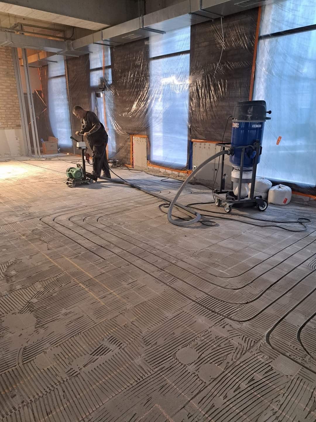 Instalacja ogrzewania podłogowego w trakcie realizacji: widoczne rury zatopione w wylewce, pracownik szlifuje powierzchnię wylewki, obok stoi odkurzacz przemysłowy; pomieszczenie w stanie surowym...