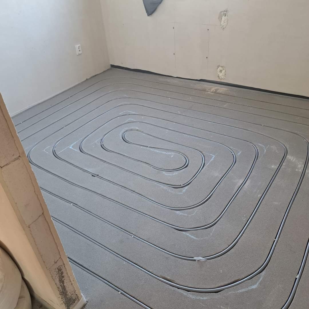 Szare rury ułożone spiralnie na podłodze z szarego styropianu, przygotowane do zalania betonem w systemie ogrzewania podłogowego.