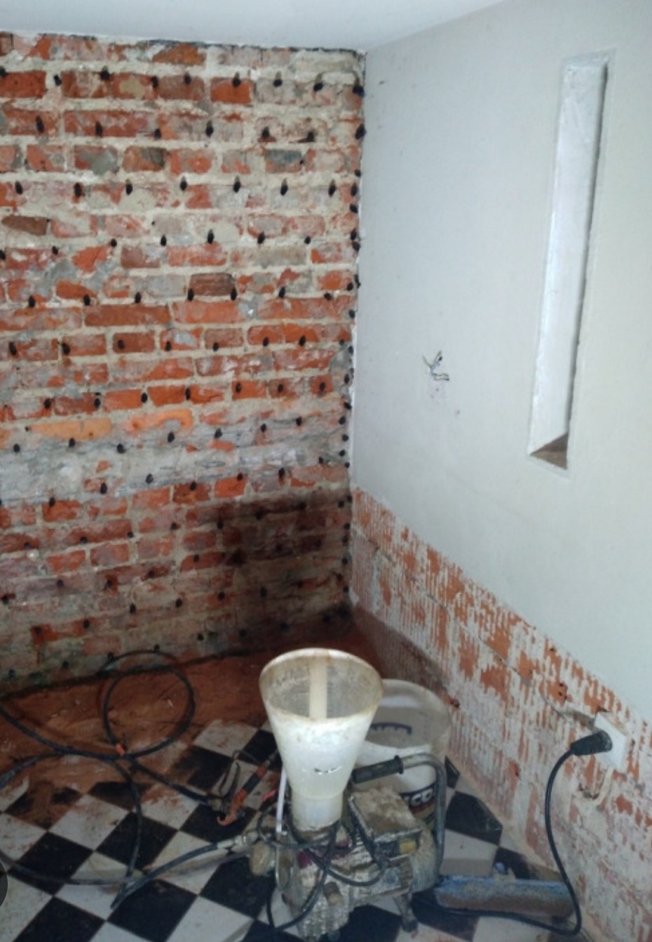 Ceglana ściana z nawierconymi otworami i wstawionymi kołkami, obok biała ściana z resztkami płytek i urządzenie do iniekcji na podłodze w kratkę.