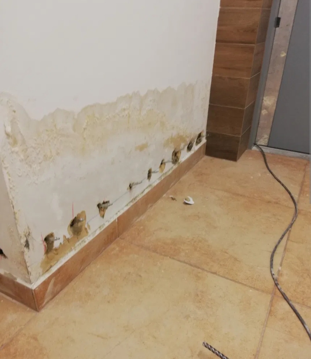 Ściana z widocznymi śladami zawilgocenia i wywierconymi otworami w dolnej części, przy podłodze wyłożonej płytkami ceramicznymi.
