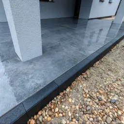 Nowoczesny taras z szarych płytek, obrzeża z czarnej kostki. Widoczne kamyki dekoracyjne. Minimalistyczny design, wysoka jakość wykonania.