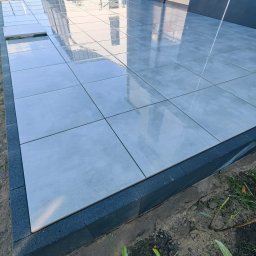 KAR-BRUK KAROL PAPROCKI - Jasnoszary taras z płytek w trakcie układania, z widoczną czarną obwódką. Kilka płytek jeszcze nie położonych. Odbicie budynku w błyszczącej powierzchni.