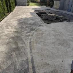 Ułożona kostka brukowa w odcieniach szarości tworzy ścieżkę z dekoracyjnymi obramowaniami. Część kostki jeszcze nieoczyszczona, w tle budynek gospodarczy i zieleń.