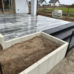 Nowoczesny taras z szaro-białej kostki, z czarnymi schodami i obrzeżem z jasnych bloczków. Częściowo ukończony, z widoczną ziemią w obrzeżu.