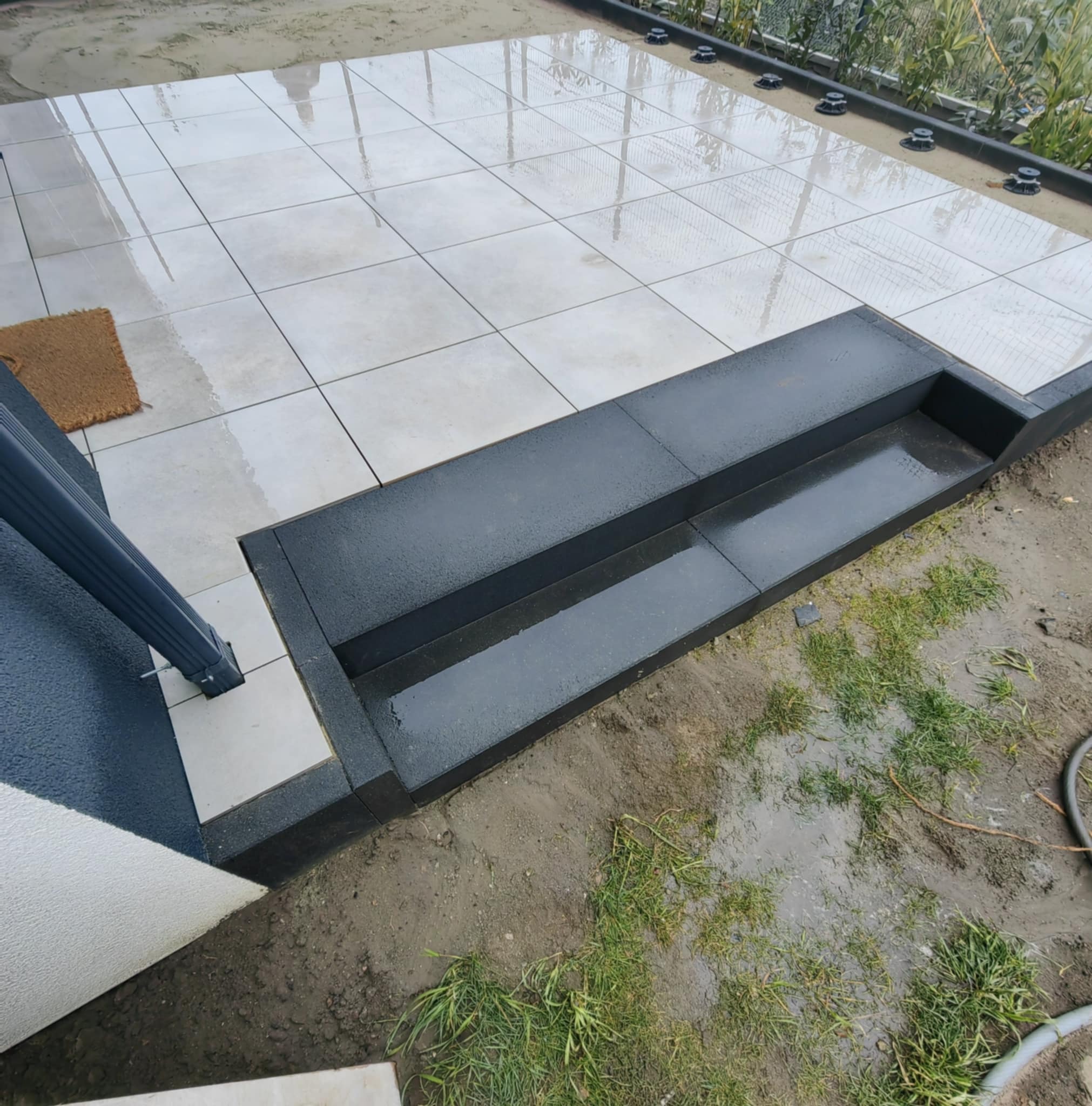 Jasne płytki tarasowe ułożone na podwyższeniu z czarnych stopni, mokre od deszczu, z widocznymi elementami montażowymi i zielenią w tle.